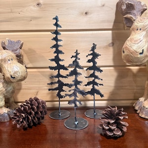 May include: Three black metal tree sculptures with circular bases stand on a wooden surface. Two pine cones and two moose sculptures flank the trees. The trees have a rustic, handmade appearance, perfect for rustic decor.