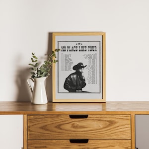 Op de afbeelding: Een ingelijste poster met een zwart-witfoto van een countryzanger met een cowboyhoed. De poster heeft de tekst "NO PLACE LIKE TOUR" bovenaan en een lijst met tourdata.