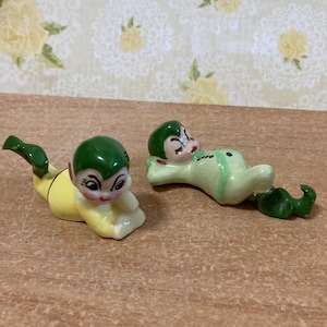 Peut inclure: Deux figurines en céramique représentant des elfes. L'un est allongé, l'autre rampe. Les elfes ont les cheveux verts, les joues roses et portent des tenues jaunes et vertes. Les figurines sont sur une surface brune.