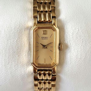 Könnte beinhalten: Eine goldfarbene Seiko Quartz Armbanduhr mit rechteckigem Zifferblatt und passendem Gliederarmband. Das Zifferblatt zeigt römische Ziffern und das Seiko Quartz Logo. Die Uhr ist ein klassisches Design, geeignet für den Alltag.