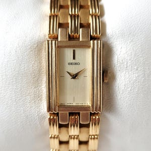 May include: A gold-tone Seiko wristwatch with a rectangular case and a matching link bracelet. The watch features a light champagne dial with simple hour markers, thin gold hands, and the Seiko logo printed in black below the twelve o'clock position.
