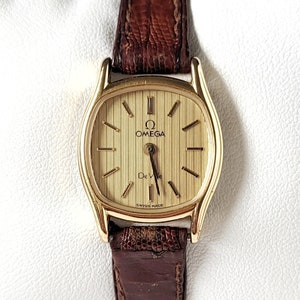 Puede incluir: Un reloj de pulsera Omega De Ville de tono dorado con una correa de cuero marrón. La esfera del reloj es rectangular y tiene una esfera de tono dorado con marcadores de hora en números romanos negros y una ventana de fecha en la posición de las 3 en punto. El reloj es de fabricación suiza.