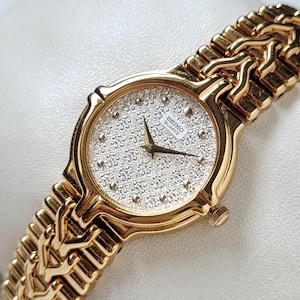 May include: A gold-tone Seiko Quartz wristwatch featuring a textured silver dial with a repeating pattern, gold-tone hands, and a polished gold-tone metal link bracelet with a distinctive interlocking design that wraps around the wrist.