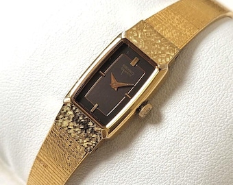 Puede incluir: Un reloj de pulsera Seiko Quartz en tono dorado con caja rectangular y esfera marrón oscuro. El reloj tiene manecillas e índices dorados, con una correa de malla metálica dorada texturizada que presenta un patrón entrecruzado cerca de la caja.