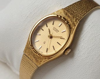 Puede incluir: Un reloj de pulsera Citizen Quartz dorado con una correa texturizada y una esfera ovalada. El reloj presenta una esfera dorada con marcadores y agujas negras. El reloj se muestra sobre una superficie blanca.