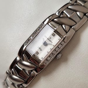 May include: A silver ROMANSON Lily Swiss Quartz wristwatch with a rectangular face. The watch face is white with a textured pattern and diamond accents. The watch band is a woven design with a clasp closure.