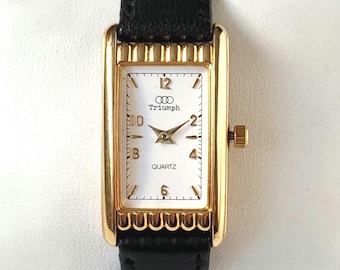 Puede incluir: Un reloj de pulsera rectangular de tono dorado con esfera blanca, manecillas doradas y números dorados en las posiciones 12, 2, 4, 6, 8 y 10. La esfera muestra el texto Triumph y QUARTZ. El reloj tiene una correa de cuero negro y un bisel dorado estriado.