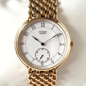 Op de afbeelding: Een goudkleurig Citizen Quartz horloge met een witte wijzerplaat en zwarte wijzers. Het horloge heeft een subdial en een goudkleurige band. De wijzerplaat toont het Citizen Quartz logo en de cijfers 1 tot en met 12.