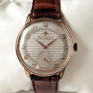 May include: A vintage Cortebert wristwatch with a silver-colored face and brown leather band. The watch face features gold-colored hands and numerals, with a small seconds dial at the bottom. The watch case is gold-toned.