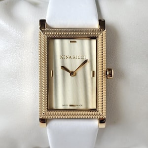 May include: A rectangular gold-tone wristwatch featuring a textured gold dial with vertical stripes, gold hands, and the text NINA RICCI and SWISS MADE. The watch has a white strap and a decorative, patterned gold-tone bezel frame.