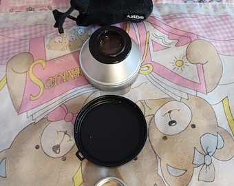 May include: A silver camera lens attachment with a black lens cap and a black drawstring bag with the word "SONY" printed on it.