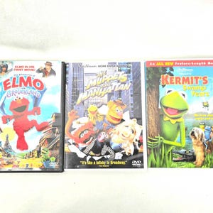 May include: Three DVD movie cases featuring colorful characters. Titles include "Elmo in Grouchland", "The Muppets Take Manhattan", and "Kermit's Swamp Years". The cases are in good condition and suitable for collectors.