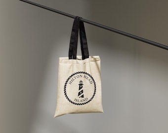 May include: A beige tote bag with black handles and a circular logo featuring a lighthouse and the text "Hilton Head Island" in a rope-style border. The bag is suspended from a black bar.