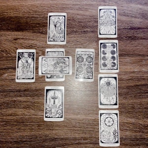 Może przedstawiać: Rozkład kart tarota z misternymi czarno-białymi ilustracjami. Karty ułożone są na drewnianej powierzchni, prezentując różne symboliczne obrazy i tekst. Układ sugeruje sesję czytania lub wróżenia.