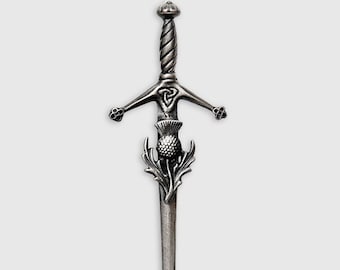 Puede incluir: Un broche de kilt plateado en forma de espada con un diseño de cardo.