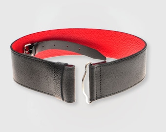 May include: A wide black belt with a red interior. The belt features a textured leather-like appearance and a silver-colored buckle closure. The belt is unfastened, showing the interior red lining.