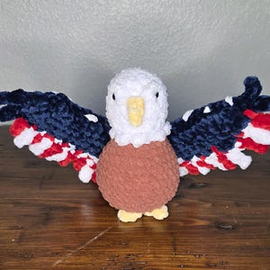 May include: A handmade crochet eagle with outstretched wings. The eagle has a white head, a brown body, and wings in navy blue, red, and white, resembling the American flag. The eagle has yellow feet and a yellow beak.