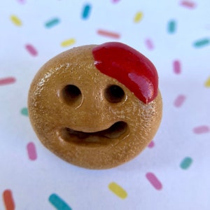 May include: A brown clay figurine with a red glaze on the top, shaped like a smiling face with two eyes and a mouth.