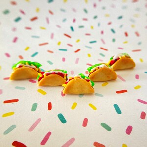 May include: Five miniature tacos made of polymer clay. The tacos are filled with red, green, and white filling and topped with white frosting. The tacos are arranged in a semi-circle on a white background with colorful confetti.