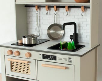 Könnte beinhalten: Eine hellgraue Spielzeugküche aus Holz mit schwarzer Armatur, Spüle und Kochfeld. Die Küche verfügt über eine Mikrowelle, einen Backofen und Stauraum. Zubehör wie Topf, Pfanne und Utensilien sind enthalten.