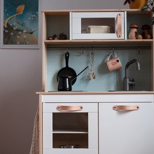 May include: A white wooden play kitchen with a black faucet, a black pot, a black pan, and a white microwave. The kitchen has a brown leather handle and a white countertop.