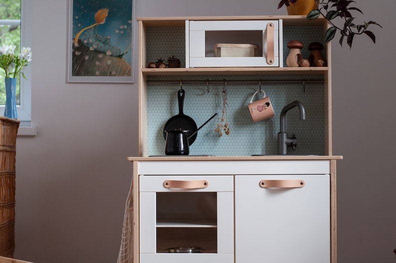 May include: A white wooden play kitchen with a black faucet, a black pot, a black pan, and a white microwave. The kitchen has a brown leather handle and a white countertop.