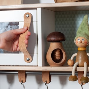 Könnte beinhalten: Ein Holzregal mit einer weißen Tür, einem Nussknacker aus Holz in Form eines Pilzes und einer Holzfigur eines Zwergs mit grünem Hut. Das Regal hat eine Metallstange mit Korkhaken.