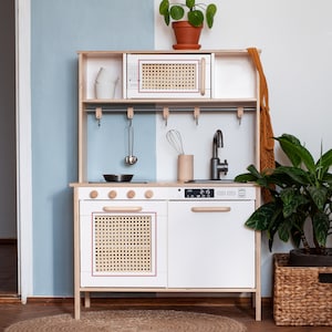 May include: A white wooden play kitchen with a black stovetop, a white oven with a woven door, a white dishwasher with a wooden handle, and a silver faucet. The kitchen has a wooden shelf with a white microwave and a woven door. There are hooks for hanging utensils on the backsplash. The kitchen is in a room with a light blue wall and a wooden floor.