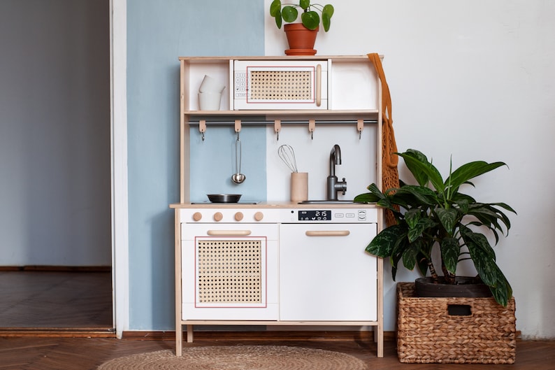 May include: A white wooden play kitchen with a black stovetop, a white oven with a woven door, a white dishwasher with a wooden handle, and a silver faucet. The kitchen has a wooden shelf with a white microwave and a woven door. There are hooks for hanging utensils on the backsplash. The kitchen is in a room with a light blue wall and a wooden floor.