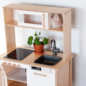 May include: A wooden play kitchen with a white microwave, a black stovetop, a grey sink, and a faucet. The kitchen has a shelf above the stovetop and a small plant in a terracotta pot.