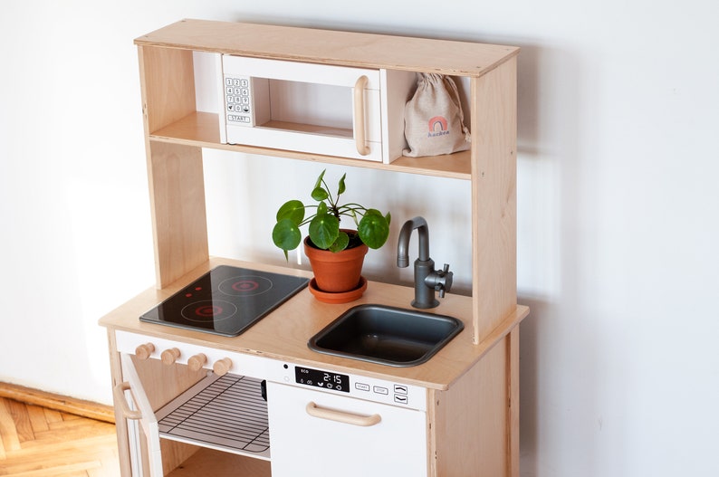 May include: A wooden play kitchen with a white microwave, a black stovetop, a grey sink, and a faucet. The kitchen has a shelf above the stovetop and a small plant in a terracotta pot.