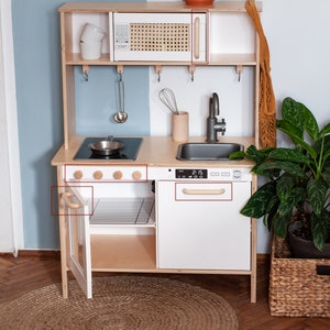 May include: A white wooden play kitchen with a black stovetop, a white sink, and a microwave with a wood-look door. The kitchen has wooden knobs and handles. The kitchen is set up in a room with a light blue wall and a wood floor.
