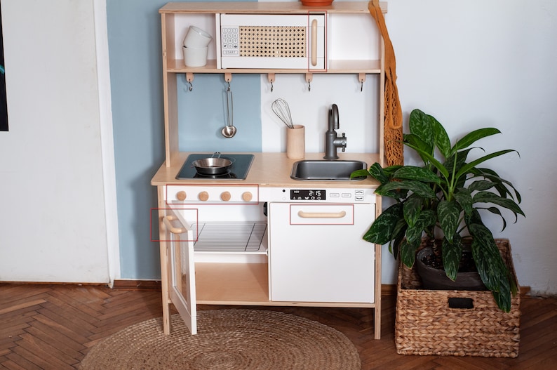 May include: A white wooden play kitchen with a black stovetop, a white sink, and a microwave with a wood-look door. The kitchen has wooden knobs and handles. The kitchen is set up in a room with a light blue wall and a wood floor.
