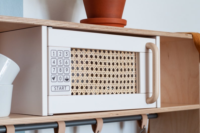 May include: White toy microwave with a wood handle and a woven rattan front panel. The microwave has a number pad with numbers 1-9 and 0, a start button, and two icons for airplane and a bowl of food.