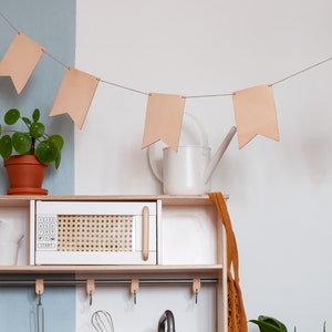 May include: A white play kitchen with a microwave, a shelf, and hooks. There is a watering can and a plant on the shelf. A string of light brown fabric flags hangs above the kitchen.
