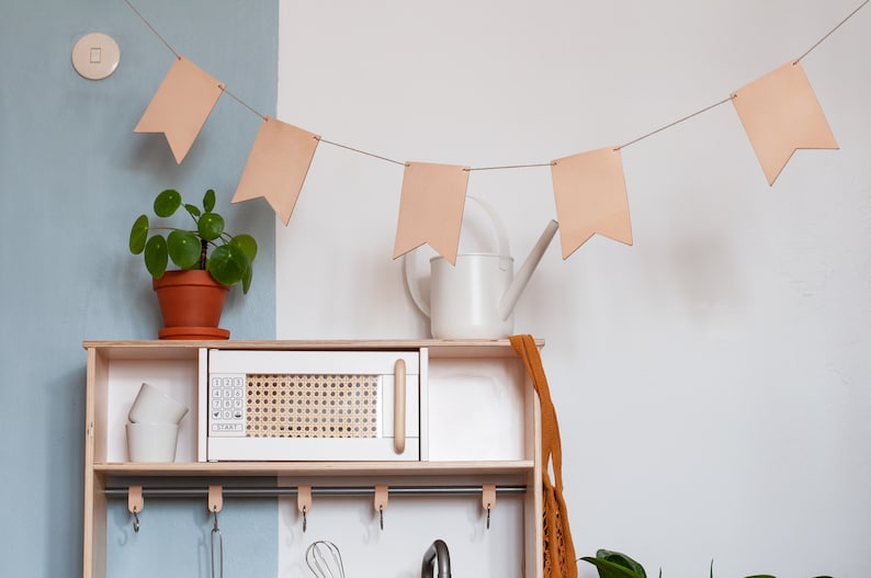 May include: A white play kitchen with a microwave, a shelf, and hooks. There is a watering can and a plant on the shelf. A string of light brown fabric flags hangs above the kitchen.