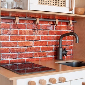 May include: A white wooden play kitchen with a black tap, a black hob, and a brick-patterned backsplash. The kitchen has a wooden shelf with hooks and a wooden worktop.