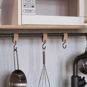 May include: A black metal bar with leather straps hangs from a wooden shelf. Five hooks are attached to the bar, and a variety of kitchen utensils are hanging from the hooks, including a whisk, a strainer, and a spatula.