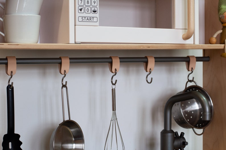 May include: A black metal bar with leather straps hangs from a wooden shelf. Five hooks are attached to the bar, and a variety of kitchen utensils are hanging from the hooks, including a whisk, a strainer, and a spatula.