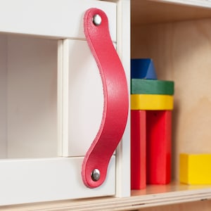 Könnte beinhalten: Ein weißer Schrank mit einem pinkfarbenen Ledergriff und silbernen Nieten. Der Schrank hat eine Glastür und ein Regal mit bunten Holzklötzen.