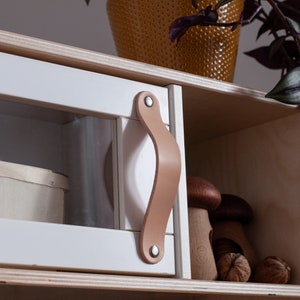 May include: A white wooden cabinet with a glass door and a leather handle. The cabinet has two shelves, one with a wooden box and the other with wooden mushrooms and nuts.
