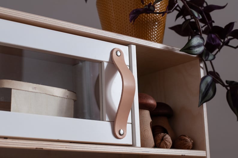 May include: A white wooden cabinet with a glass door and a leather handle. The cabinet has two shelves, one with a wooden box and the other with wooden mushrooms and nuts.