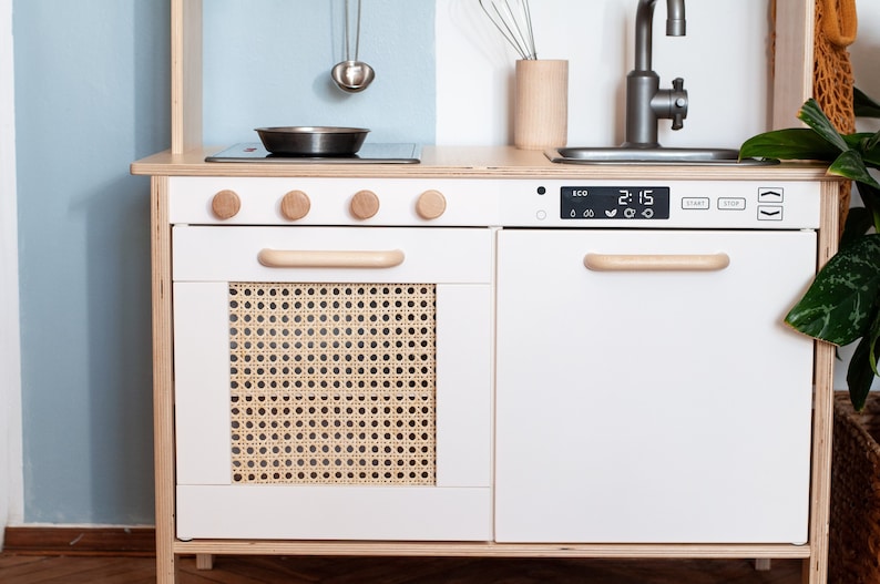 May include: White wooden play kitchen with a stove, oven, and sink. The oven door has a woven rattan panel. The stove has three knobs and a digital timer that reads "2:15".