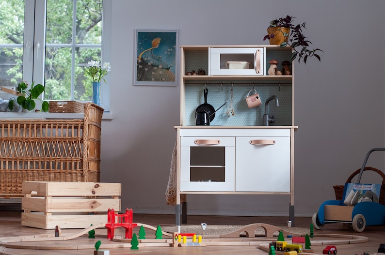 May include: A white wooden play kitchen with a black stovetop, a silver faucet, and a brown leather handle. The kitchen has a small oven, a shelf, and a hanging rack. There are wooden train tracks and a red bridge on the floor in front of the kitchen.