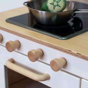 May include: A white play kitchen with a wooden countertop and a black electric stovetop. The stovetop has a stainless steel pot with a green star-shaped toy on top. The kitchen has four wooden knobs and a wooden handle on the oven door.