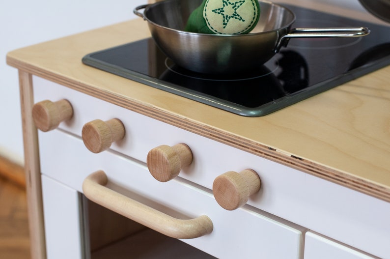 May include: A white play kitchen with a wooden countertop and a black electric stovetop. The stovetop has a stainless steel pot with a green star-shaped toy on top. The kitchen has four wooden knobs and a wooden handle on the oven door.