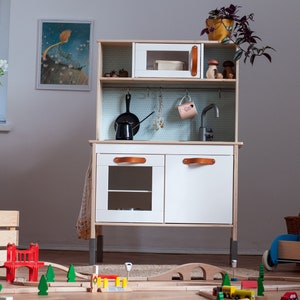May include: A white wooden play kitchen with a black sink, a microwave, and two cabinets with brown leather handles. The kitchen is on a wooden floor with a toy train track and a red bridge.