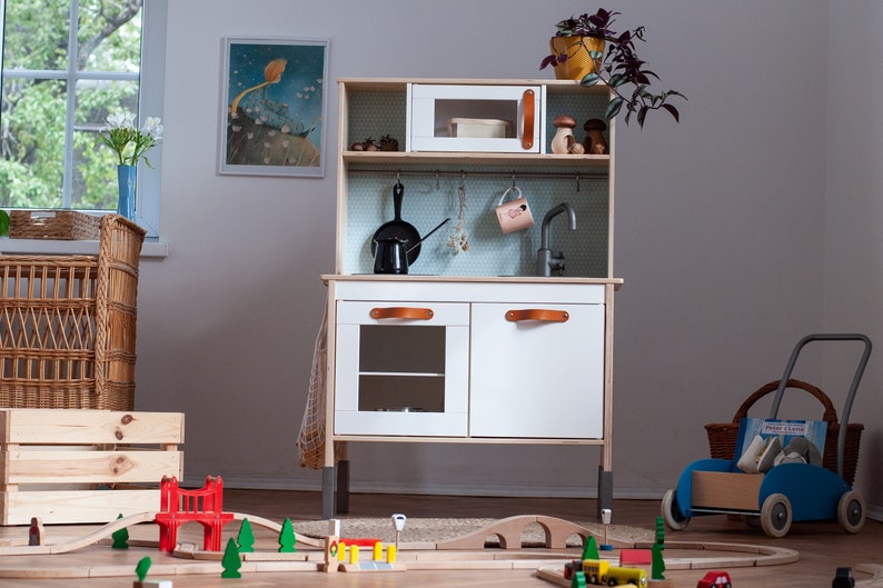 May include: A white wooden play kitchen with a black sink, a microwave, and two cabinets with brown leather handles. The kitchen is on a wooden floor with a toy train track and a red bridge.