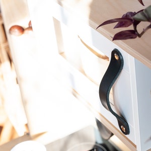 May include: White wooden box with a black leather handle and gold metal accents. The box is part of a set of shelves.