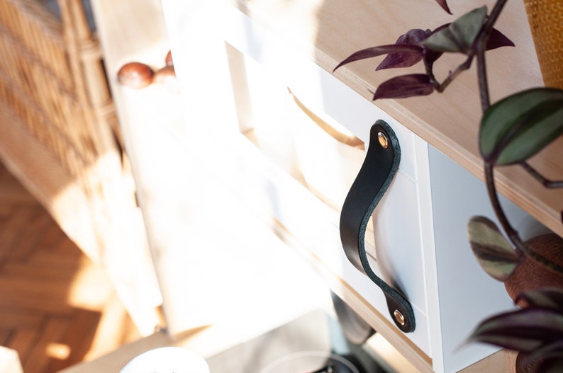 May include: White wooden box with a black leather handle and gold metal accents. The box is part of a set of shelves.